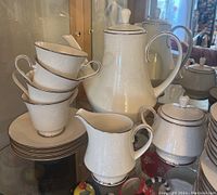 Photo of stacked teacups, saucers, coffee pot, creamer, and sugar bowl showing embossed floral pattern with silver rims.