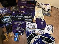 Wide view of Colorado Rockies apparel laid out on carpet, showing various shirts, hoodies, jerseys, hat, scarf, and memorabilia items.