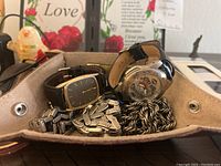 Close-up photo of a small beige fabric tray holding various jewelry and watches including a rectangular gold-tone wristwatch, a sports-themed watch featuring a Super Bowl Champions logo, metal cufflinks, and a thick chain.