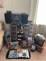 Wide view of stereo system with dual speakers on top of a stand surrounded by stacks of CDs and cassette tapes.