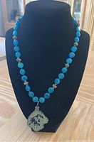 Turquoise blue and gray bead necklace with rough crystal geode pendant displayed on black jewelry bust.