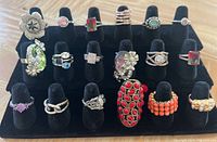 Photo showing 16 rings displayed on a black velvet ring stand, various styles and colors including red, orange, purple, green and clear stones