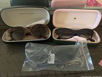 Two pairs of sunglasses side by side in their open branded cases plus a third pair sealed in plastic bag.