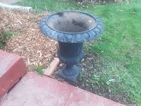 Photo showing black cast iron planter urn with embossed petal design on the rim and pedestal base, placed next to garden bed and red stone walkway. Shows some outdoor wear and rust inside.