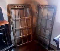 Photo of two old wooden farmhouse windows leaning against a wall, showing the full front view of both windows, one missing some glass panes