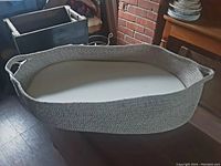 Oval rope pet bed with high sides and handles, shown with oval cushion inside on floor by brick wall.