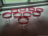 Nine ruby glass dessert compotes arranged in two rows on a white surface next to a window, showing clear glass bowls with ruby red rims and bases.
