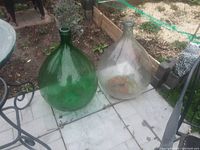 Two demi john bottles placed on light-colored patio tiles outdoors, one green and one clear glass, showing overall shape and size