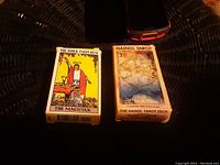 Two tarot card decks in their original boxes placed on a dark textured surface. The Rider Tarot Deck box features The Magician card. The Haindl Tarot Deck shows an artistic, muted color design.