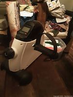 Side angled view of white under desk stationary bike showing pedal with strap, resistance knob, and digital display mounted on the top