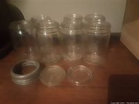Four clear glass jars and two glass lids with one metal lid displayed separately to show accessories included.