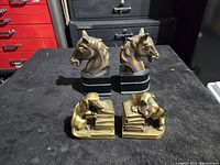 Two sets of vintage bookends placed on a black surface in front of black drawers, one set with horse heads and one set with dog reading figurines on books.