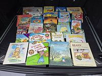 Wide shot of all 28 assorted vintage children's books spread on black surface showing colorful covers including Emma's Pet, If You Give a Mouse a Cookie, Beany, Winnie the Pooh, and more.