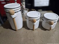 Full set of 3 white ceramic canisters with attached wooden spoons of varying sizes on a wooden surface