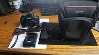 Canon EOS Rebel T3 camera body, two battery chargers, Canon branded camera strap, power adapter, and Sony branded soft camera carrying case on wooden desk.