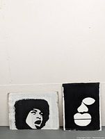 Two original paintings on recycled cardboard leaning against wall. One is a black and white portrait with afro hair, the other is a minimalist abstract face painting.