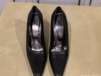 Top-down view of pair of black Calvin Klein pumps with pointed toes and moderate heels on a beige flat surface.