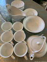 Photo showing white ceramic teapot, six white cups, clear decorative glass, Corelle plates and bowls, and a white ceramic gravy boat all arranged on table.