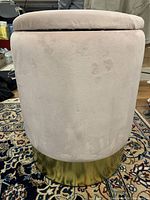 Side view of round beige fabric storage ottoman with gold base, showing some wear and marks.