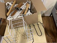 Box containing assorted white plastic and wooden clothing hangers with metal clips, two black metal hangers, and one tall white metal tiered hanger on a wooden floor and table.