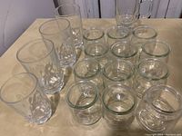 Photo of 5 tall drinking glasses with textured bottoms and 10 small clear glass jars on a table.