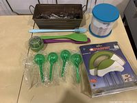 Overview of brown cutlery tray holding assorted silverware, two silicone spatulas, small cleaning brush, green watering bubbles, and boxed Starfrit burger patty maker with blue lid container.