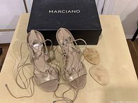 Pair of Marciano heeled sandals with ankle wrap ties and metallic silver heels placed next to the original box.
