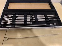 Full view of stainless steel fork and cutting utensil set in black carrying case with slots and loops, with one metal skewer pulled out on table