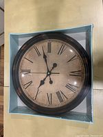 Photo showing the large round wall clock in its sealed packaging with black Roman numerals and dark frame.