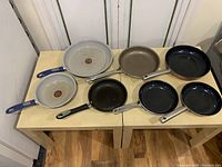 Overview of 7 frying pans of various sizes and materials arranged on a wooden surface, used condition with visible scratches