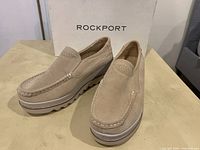 Pair of beige suede Rockport slip-on shoes displayed outside box, showing profile and top with moc toe stitching and perforations.