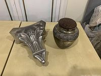 Metallic urn with patterned brown lid and silver ornamental band next to silver decorative wall sconce on a table.