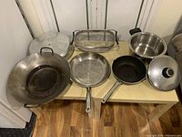 Photo showing the entire lot of kitchenware including metal wok with base and lid, multiple pans, stainless steel pot, and glass baking dish arranged on a table.