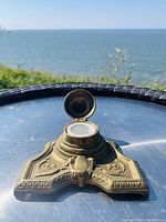 Brass triangular inkwell with lid open showing white ceramic ink reservoir inside, set outdoors on metal surface with ocean backdrop.