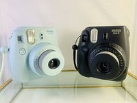 Front view of light blue and black Fujifilm Instax Mini 8 cameras side-by-side, showing lens, flash, and focus dial