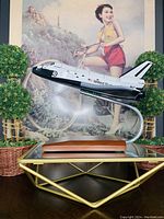Model of the Space Shuttle Discovery mounted on clear curved stand with wooden base, situated on a table in front of artwork and decorative plants.