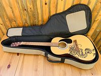 Full view of the acoustic guitar in the open soft case resting on wooden surface. Guitar has natural wood finish and dragon design on the body.