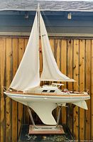 Front and side view of the vintage wooden sailboat model with fabric sails mounted on a metal and wood stand against a wooden fence background.