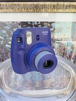 Front-left angle showing the lens extended, flash, lens markings, and shutter button on the purple Instax Mini 8 camera.