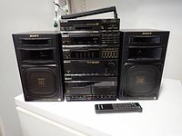 Front view of Sony audio system with two speakers on sides and remote controller in front. System stacked with audio components: DVD player, equalizer, cassette deck, tuner, amplifier. All in black with gold Sony branding.