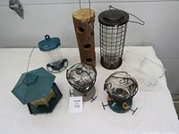 All seven bird feeders displayed on a flat surface showing different types including glass hummingbird feeders, plastic window feeders, suet feeder, wooden log feeder, and seed feeder.
