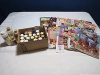 Wide shot showing a box of various acrylic paint bottles and jars, several craft magazines and books spread out on the table including titles related to tole painting and seasonal crafts.
