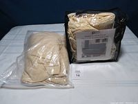 Two packaged beige stretch furniture covers for sofa and loveseat, one in sealed plastic with label showing fit information, the other in a black zippered bag with label showing sofa image and text
