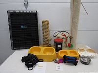 Photo showing the pet crate, two scratching posts, feeding dishes, cat harness, leash, toys, and treat container all arranged on a white surface with a plain background.