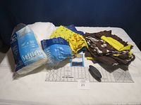 Photo of the sewing lot showing two bags of SoftLoft polyester batting, assorted fabric pieces in yellow, blue, and brown, O'Lipfa no-slip ruler, and sewing accessories on white surface.
