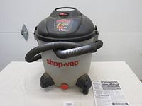 Front view of the QSP Shop-Vac Plus 10 gallon wet/dry vacuum with flexible hose wrapped around top, red Shop-Vac branding visible on the tank, and instruction sheet laid next to it.