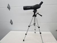 Side view of black Meade spotting scope mounted on aluminum tripod extended fully on a white table against a white wall.