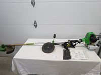 Full view of the trimmer/brush cutter laid flat on white cloth surface, showing the green engine at one end, long silver shaft, circular metal cutter blade mid-shaft, string trimming head opposite end, and parts list manual alongside