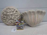 Front view of sun god plaque, fountain basin, and boxed Lagun water pump placed on white surface against neutral background.