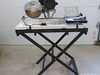 Full view of the wet tile cutting saw mounted on an X-frame metal stand indoors with garage door in background. Black and white color tray beneath blade.